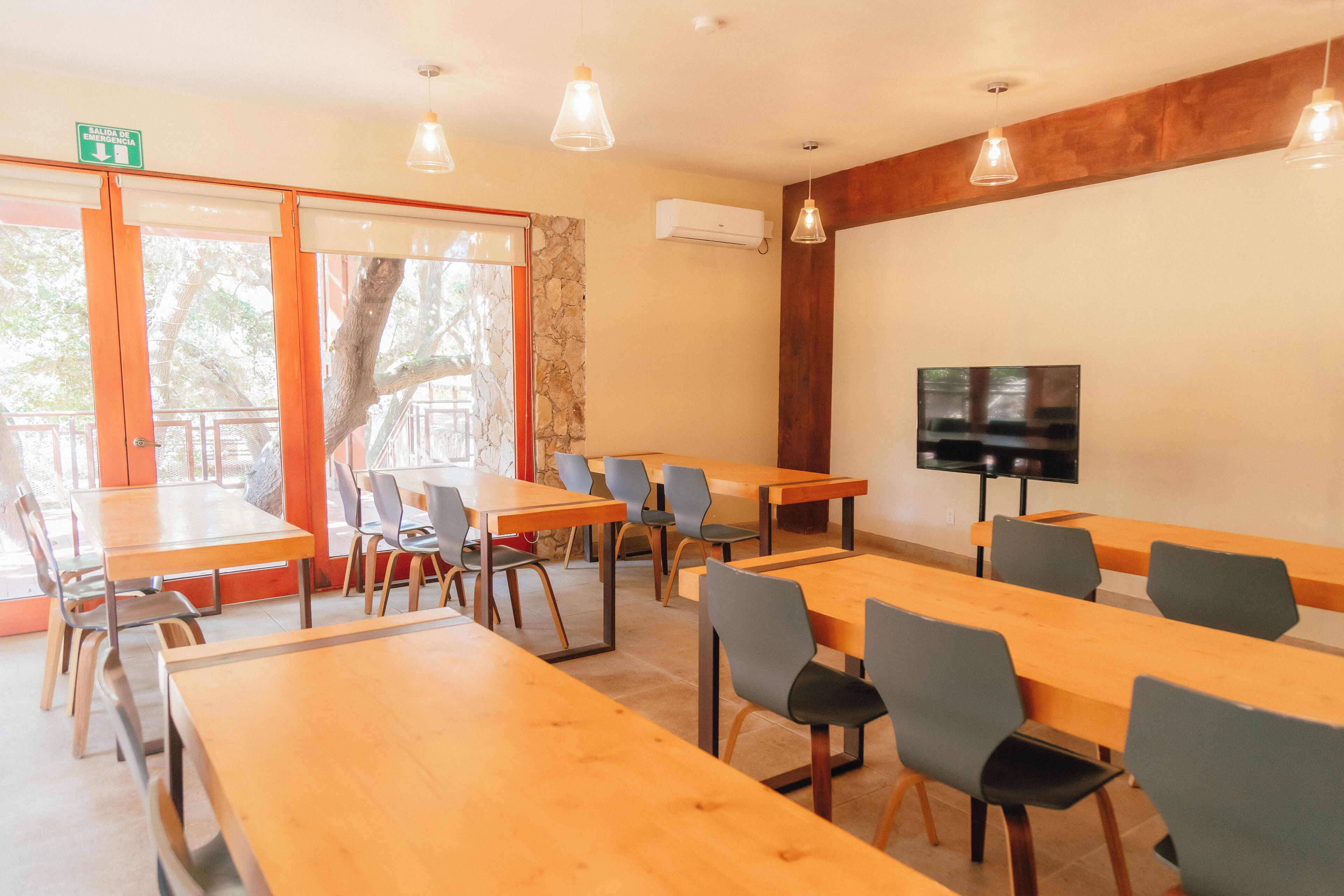 Ein Konferenzraum mit Holztischen und -stühlen, einem Fernseher an der Wand und einem großen Fenster mit Blick auf einen Baum.