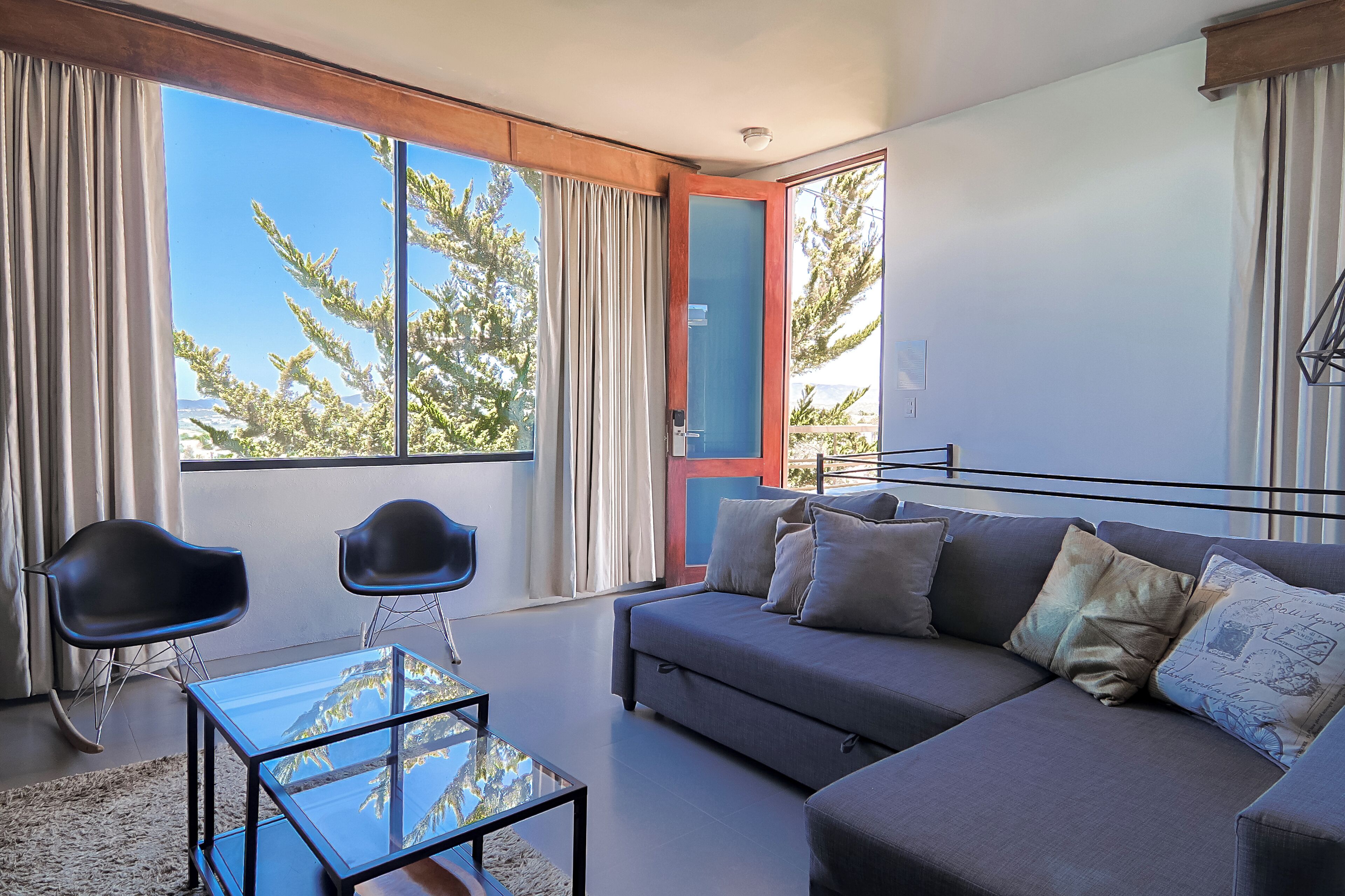 A modern living room with a sofa, two chairs, a glass coffee table, and a large window with curtains.