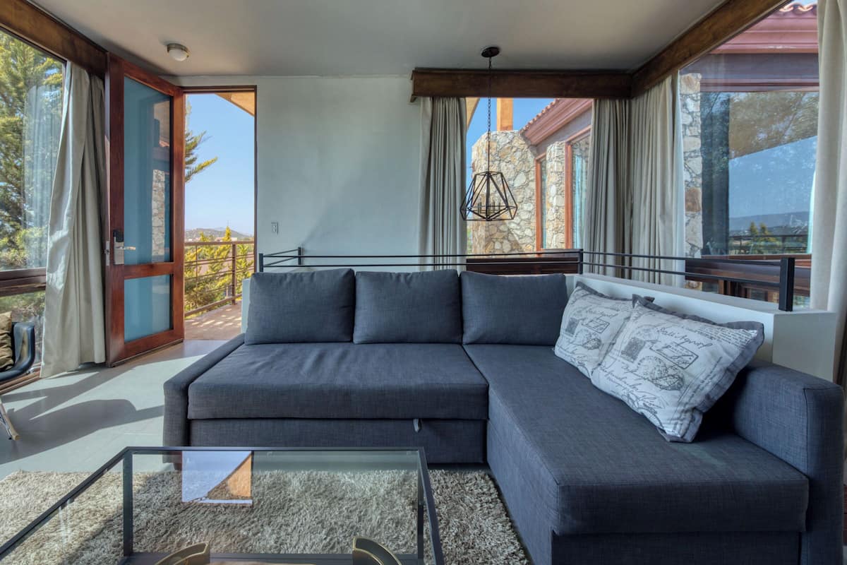 Ein modernes Wohnzimmer mit einem grauen Ecksofa, einem Glastisch und Blick ins Freie durch große Fenster.