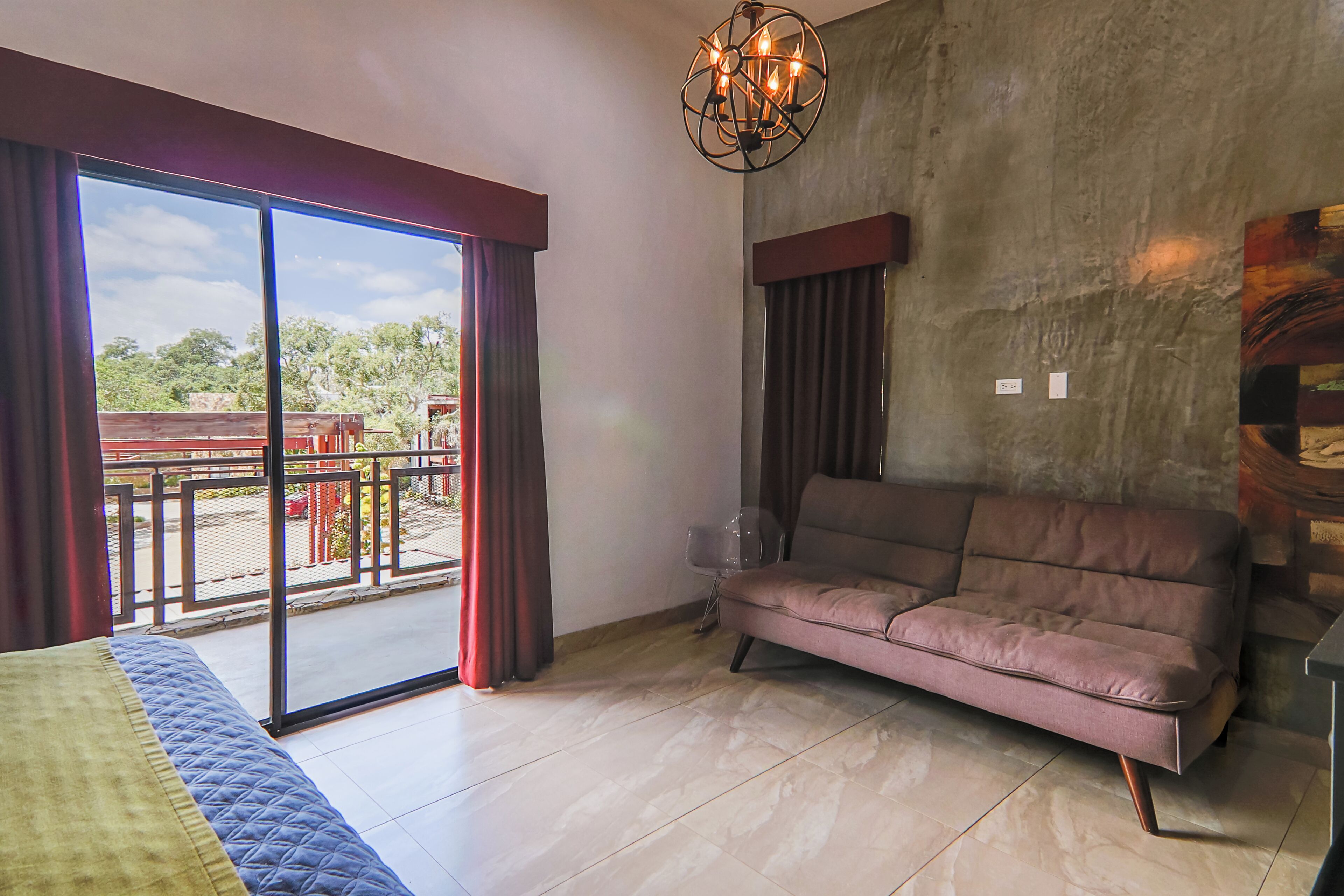 A room with a bed, a sofa, a large window with curtains, a chandelier, and a view of the outside.