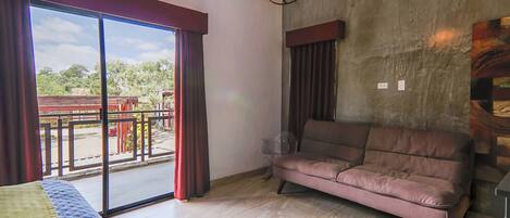 A room with a bed, a sofa, a large window with curtains, a chandelier, and a view of the outside.