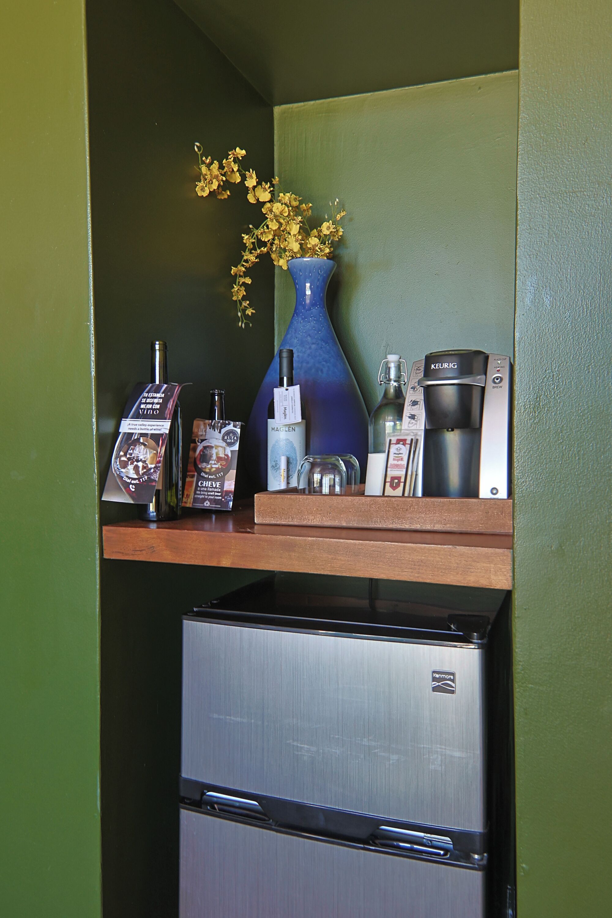 A kitchenette with a coffee maker, a bottle, and a vase with dried flowers on a shelf.