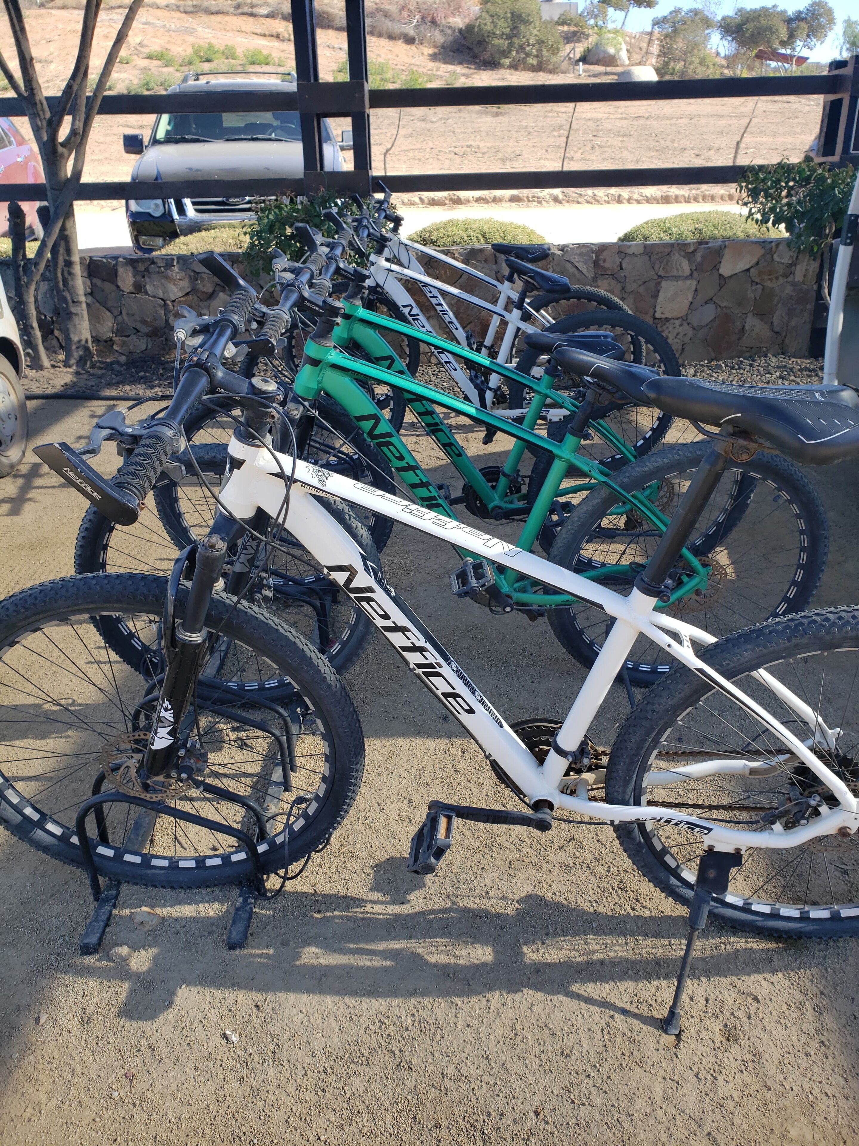 Una fila di biciclette parcheggiate all'esterno, con un muro di pietra e un'auto sullo sfondo.
