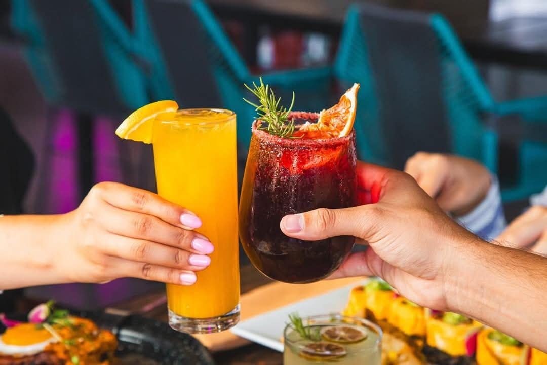 Two people holding glasses of beverages, one orange and one dark red, with garnishes.