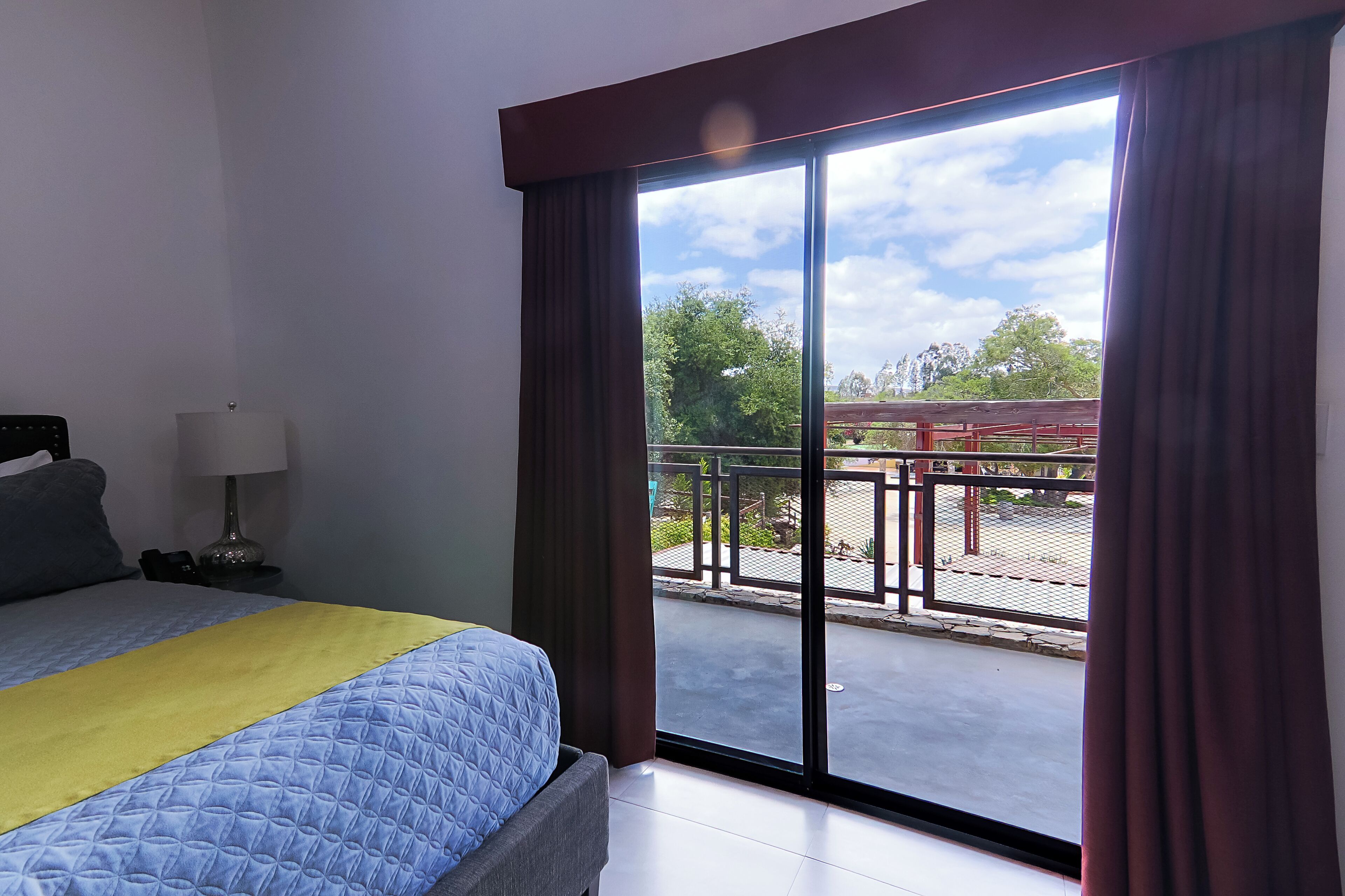 Habitación de hotel con cama, mesita de noche con lámpara y vista al balcón exterior.