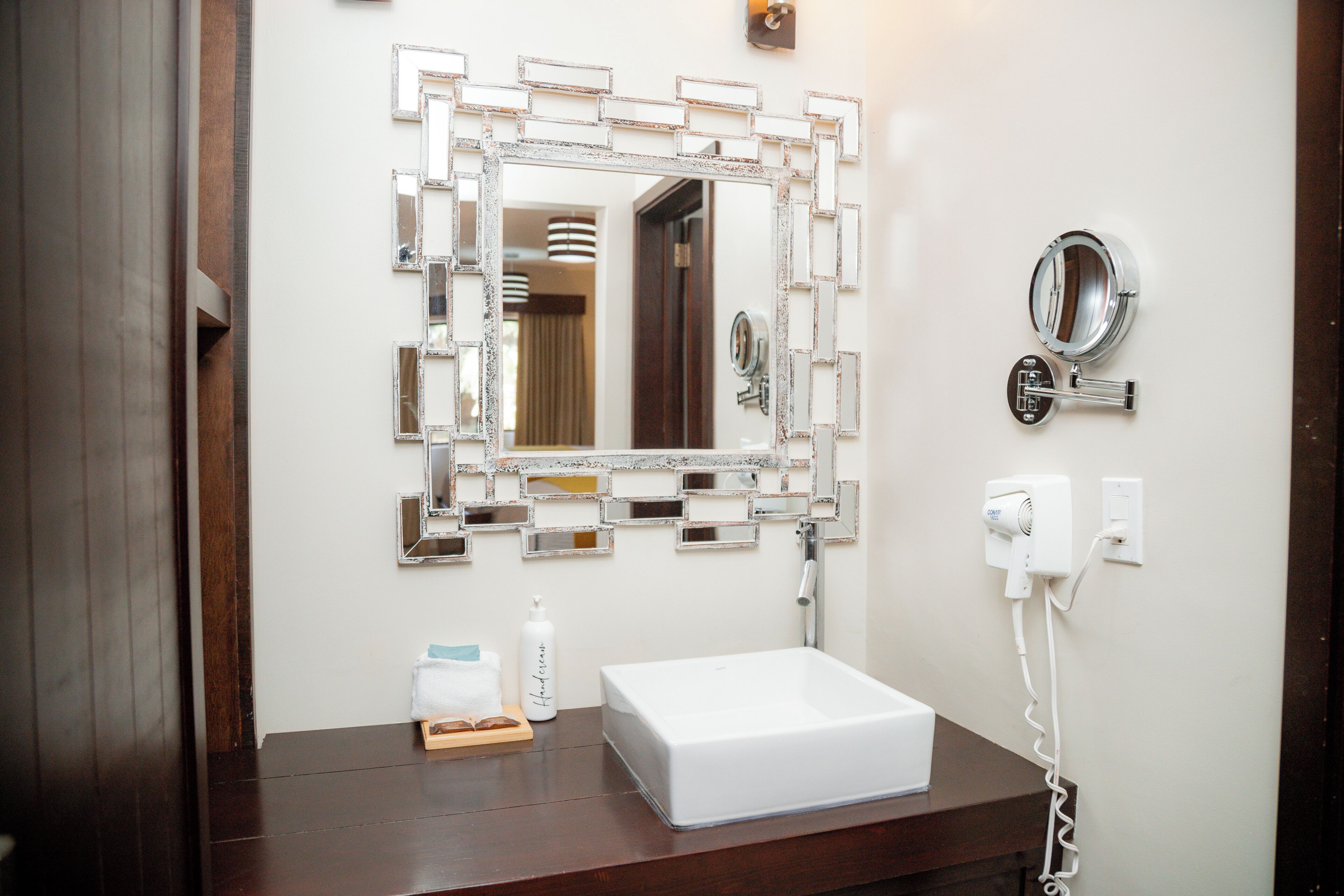 A bathroom with a large mirror, a white sink, a handheld mirror, and a hairdryer on the wall.