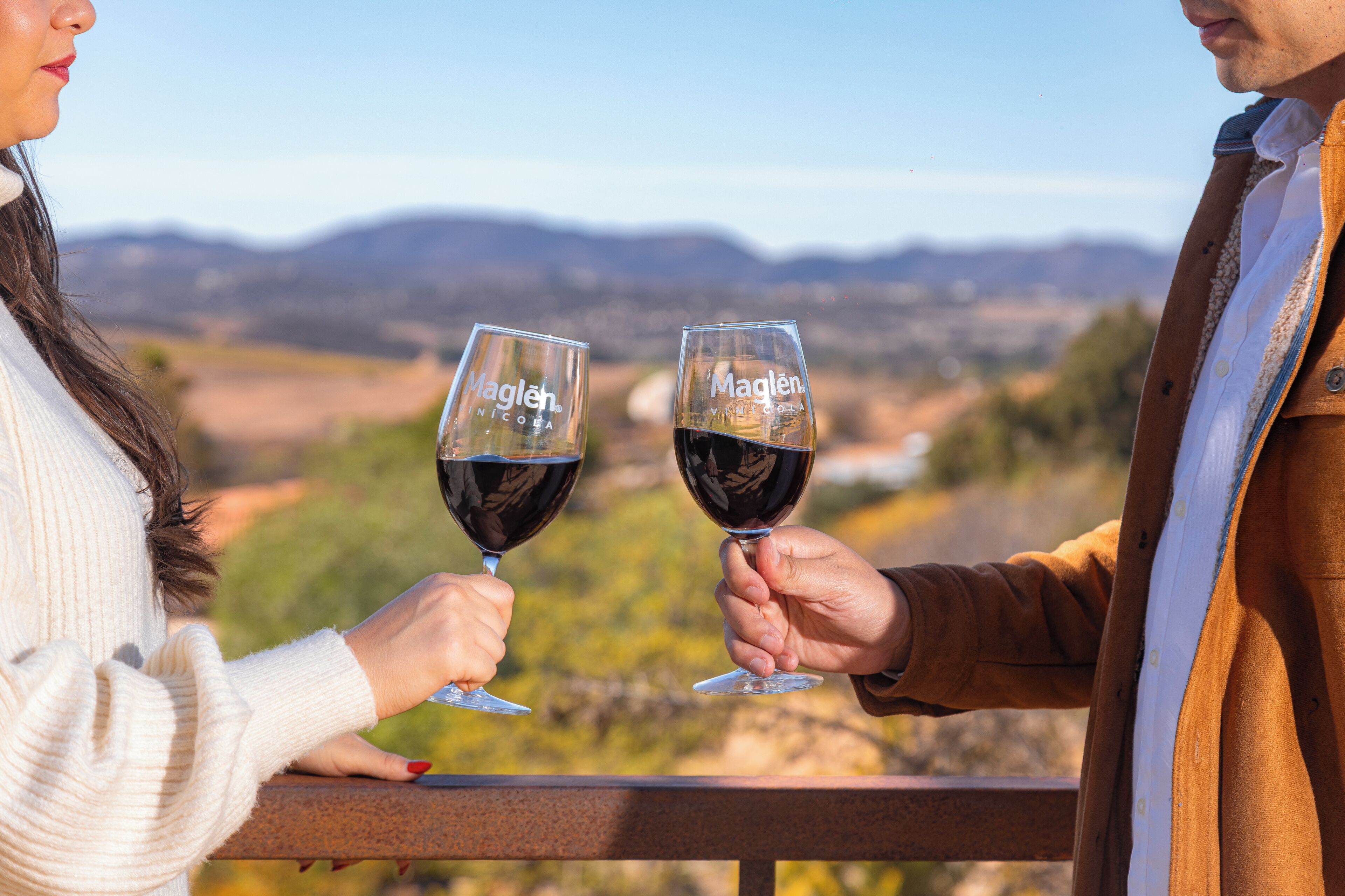 Two people holding wine glasses with 'Maglen' on them, overlooking a scenic landscape.