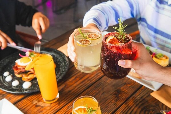 A person holding two drinks with garnishes, a plate with food, and a glass of orange juice on a wooden table.