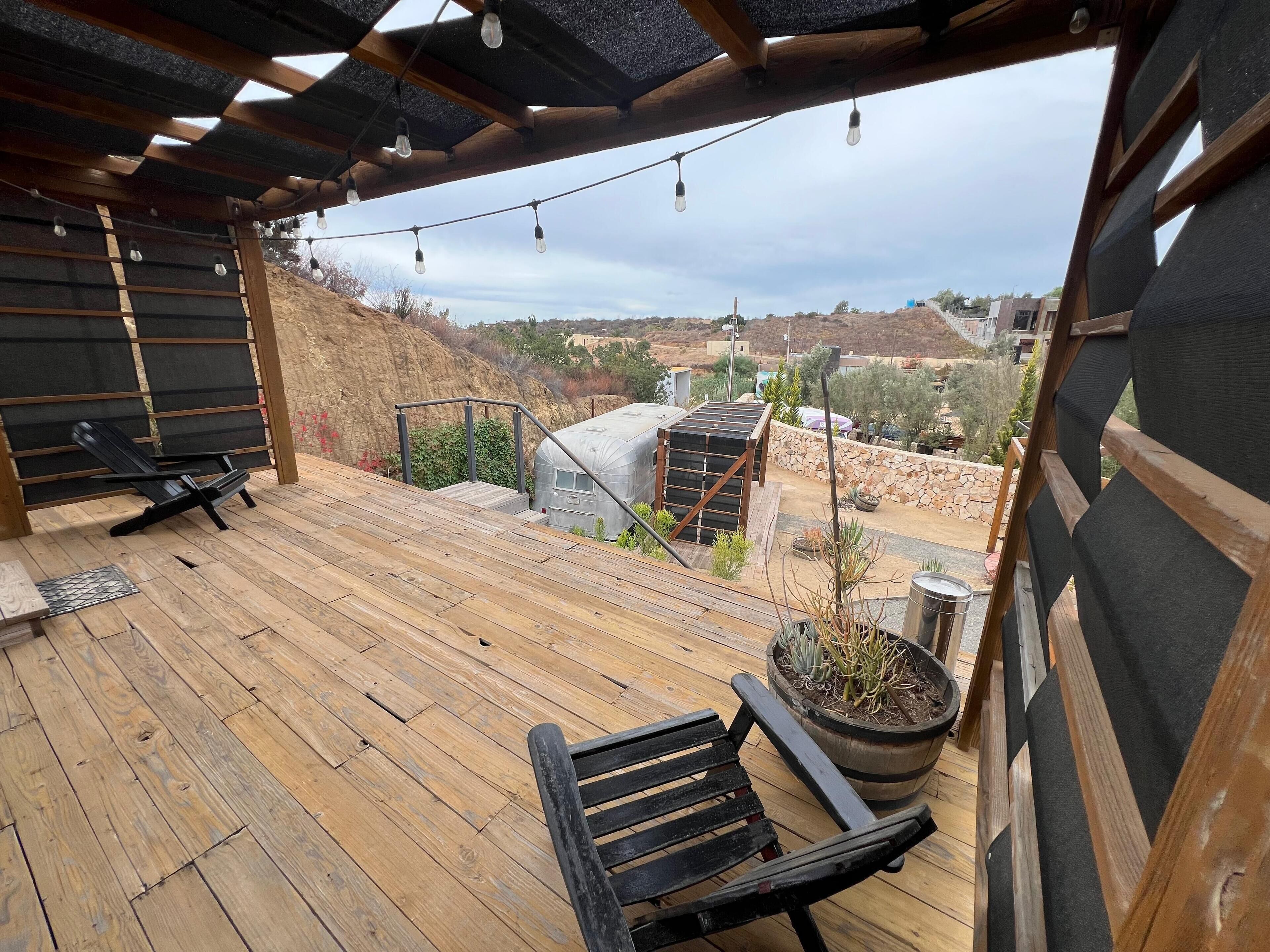 A wooden deck with a bench and a potted plant, overlooking a hilly landscape with buildings in the distance.