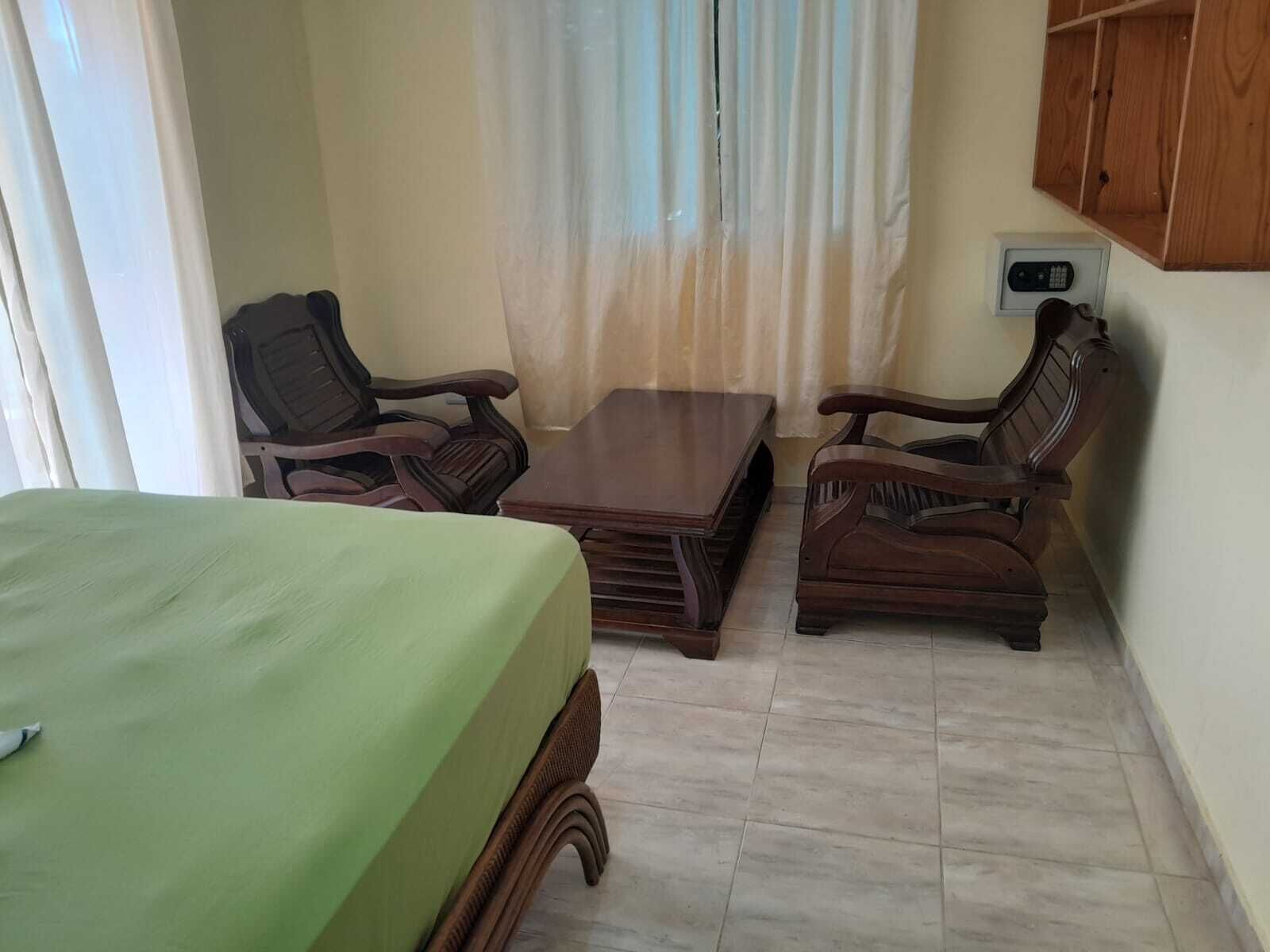 Una habitación con una cama, dos sillas de madera, una mesita pequeña y un armario de madera.