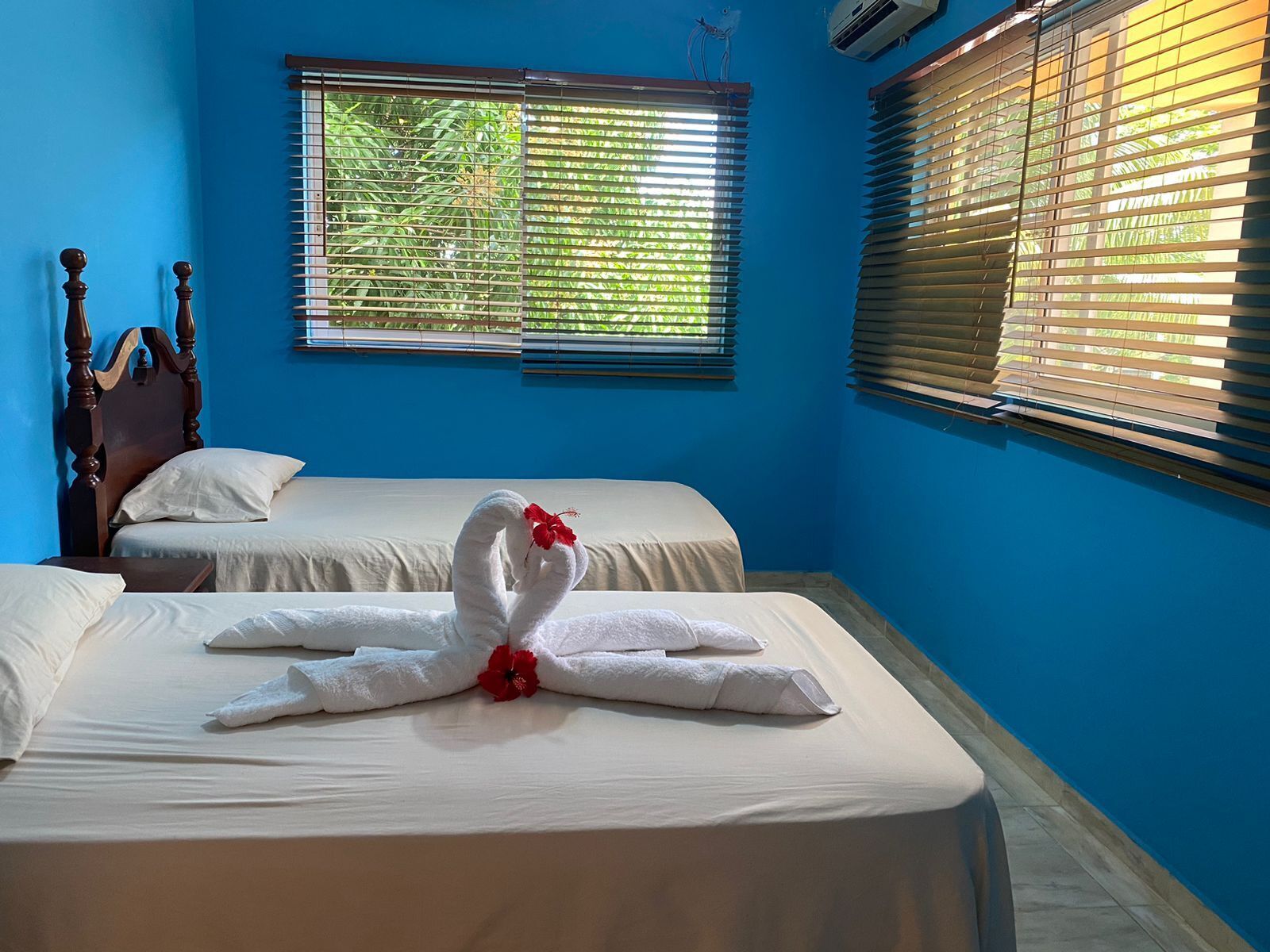 A hotel room with two beds, a wooden headboard, a window with blinds, and a red hibiscus flower on the bed.