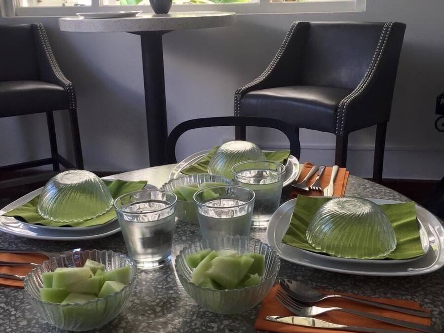 A dining table set with clear glasses, a bowl of green fruit, and a potted plant in the background.