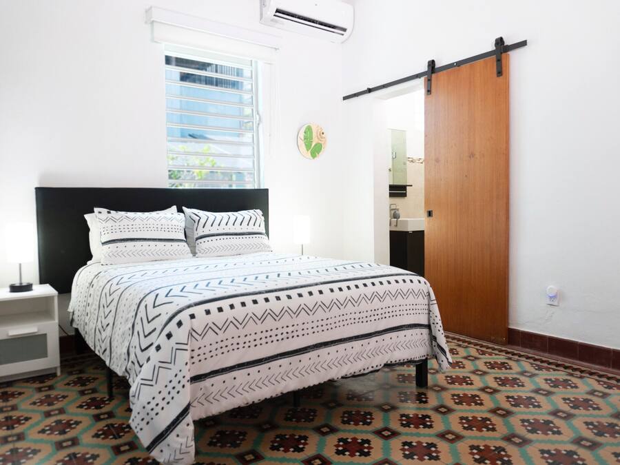 Un dormitorio con cama, mesita de noche, ventana, puerta y suelo estampado.