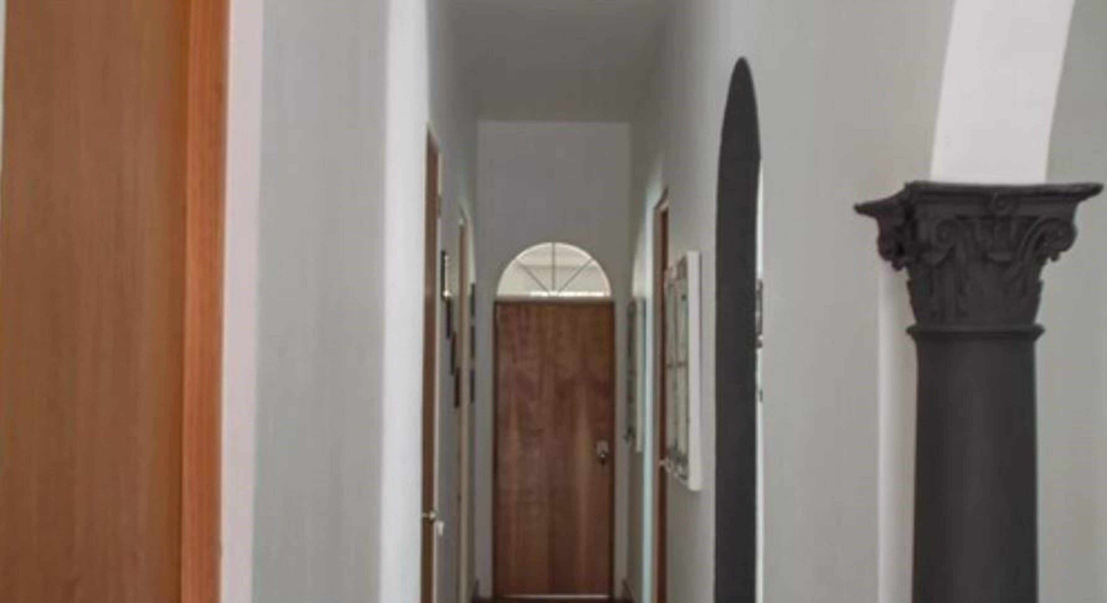 A hallway with a wooden door at the end, a black column with ornate detailing, and a white wall with a window.