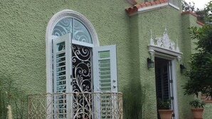 A green stucco building with a white door and window shutters, a small balcony with ornate metal railings, and a decorative pediment above the door. - Casa Isabel Bed & Breakfast (San Juan)