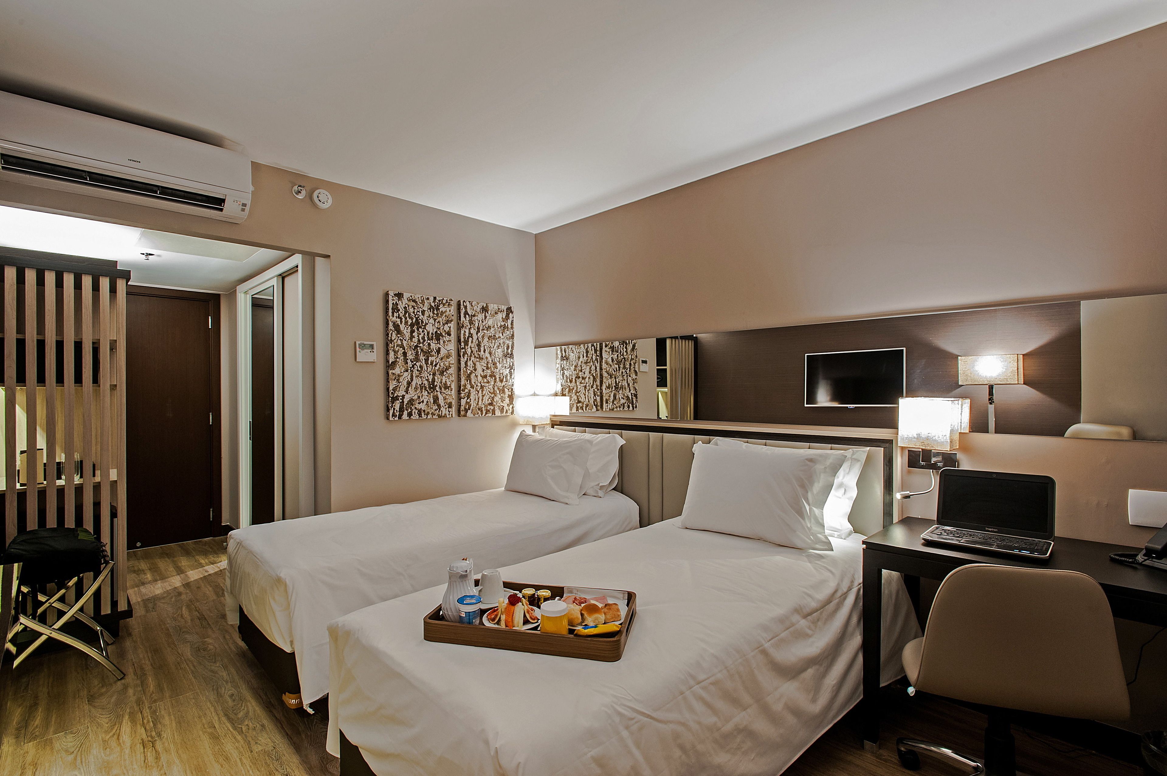 A hotel room with two beds, a desk with a laptop, a tray of breakfast items, and a TV on the wall.
