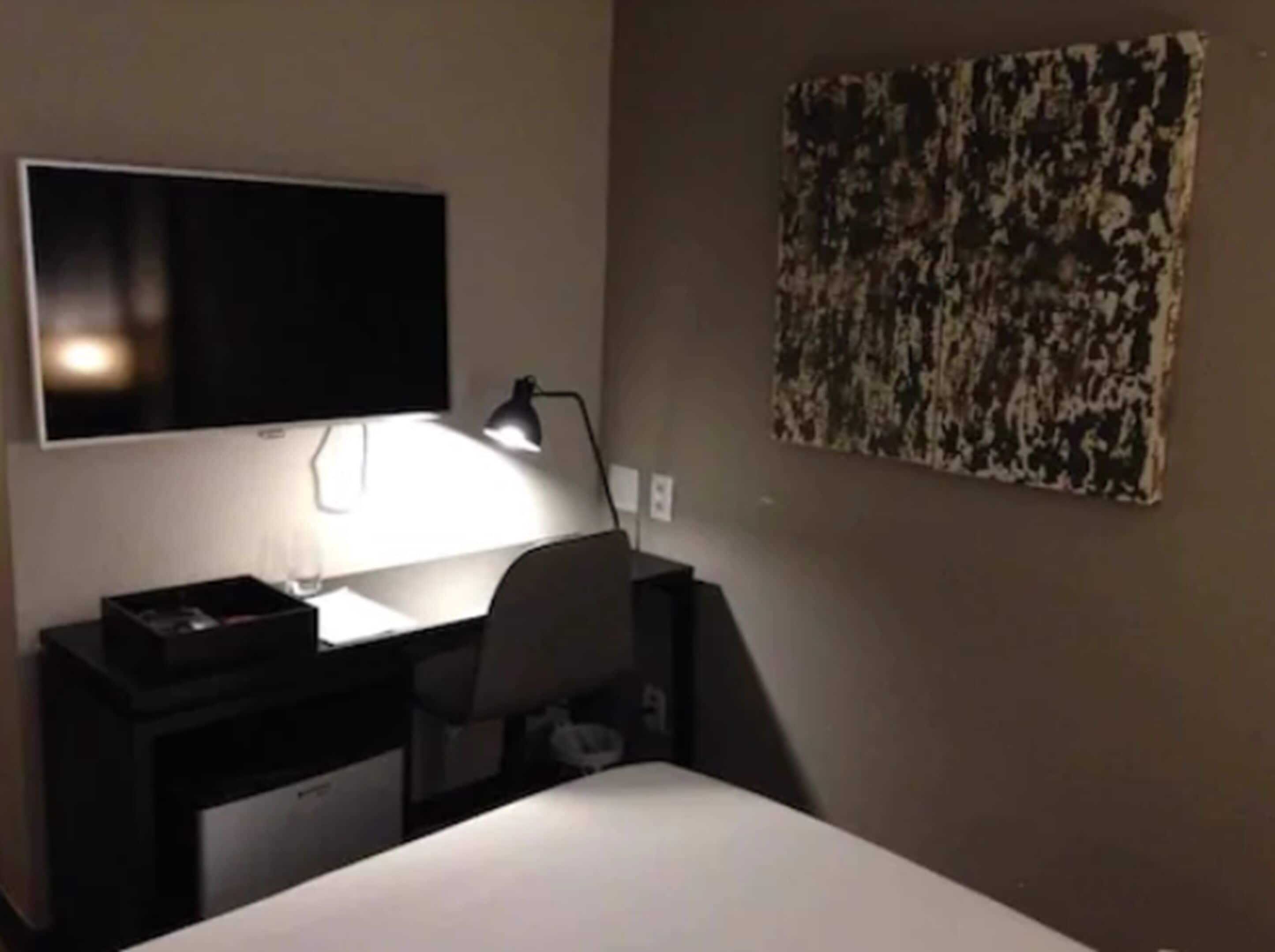 A hotel room with a bed, a desk with a lamp, a flat-screen TV, and a painting on the wall.