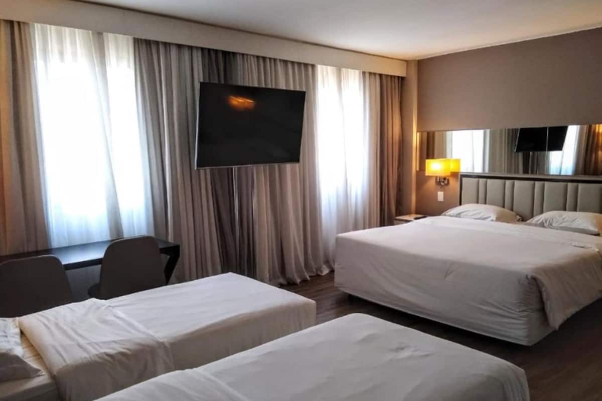Quarto de hotel com duas camas, uma escrivaninha, uma TV e cortinas.
