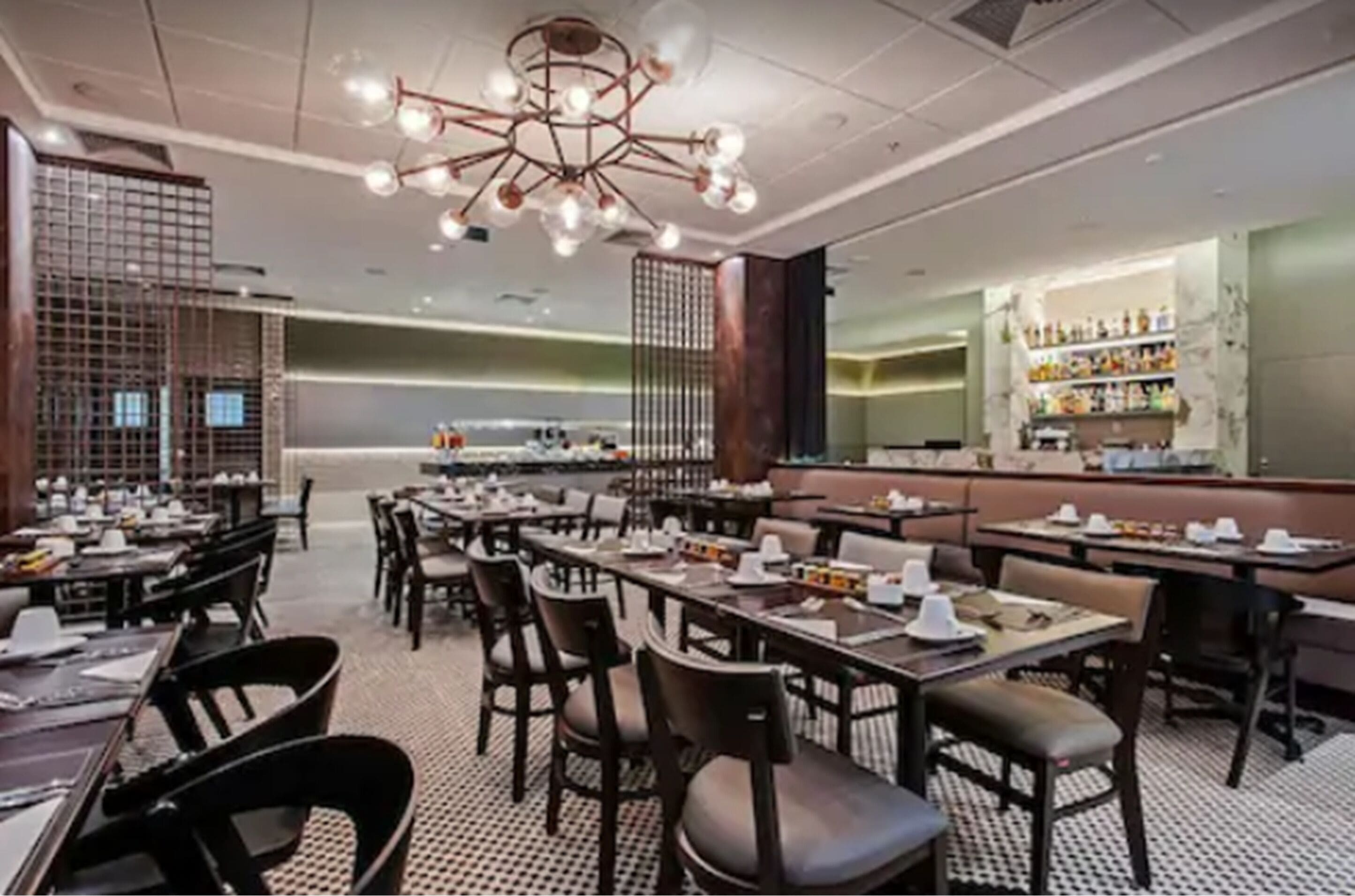 A restaurant interior with multiple tables and chairs, a central chandelier, and a bar area with shelves of bottles.
