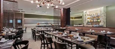 A restaurant interior with multiple tables and chairs, a central chandelier, and a bar area with shelves of bottles.
