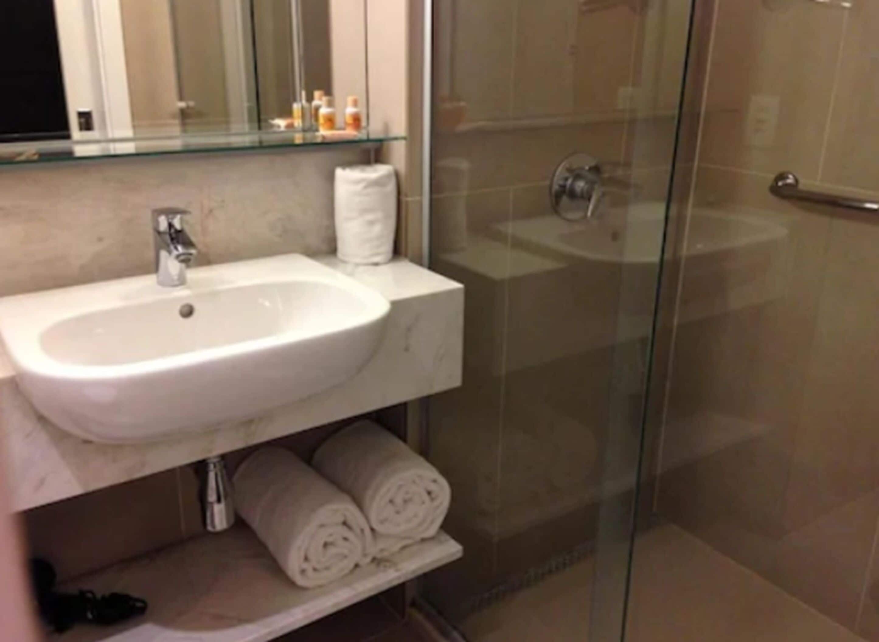 A bathroom with a white sink, a glass shower enclosure, and a shelf with rolled towels.