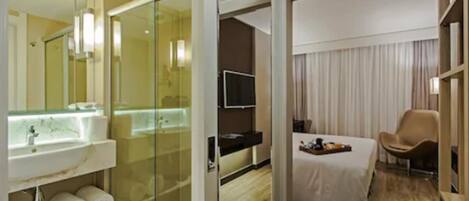 A hotel room with a glass shower enclosure, a bed with a white bedspread, a television, and a chair.