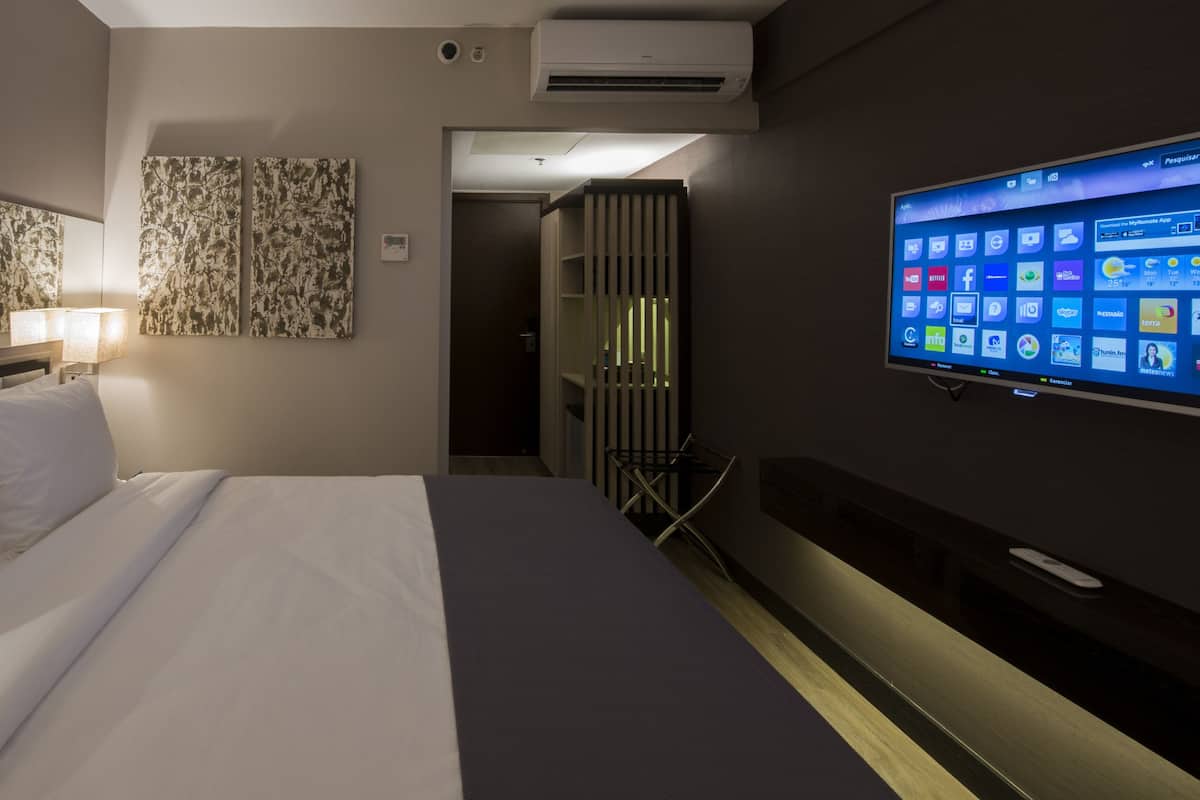 Quarto de hotel com uma cama, uma televisão na parede exibindo a tela inicial e um espelho sobre a cama.