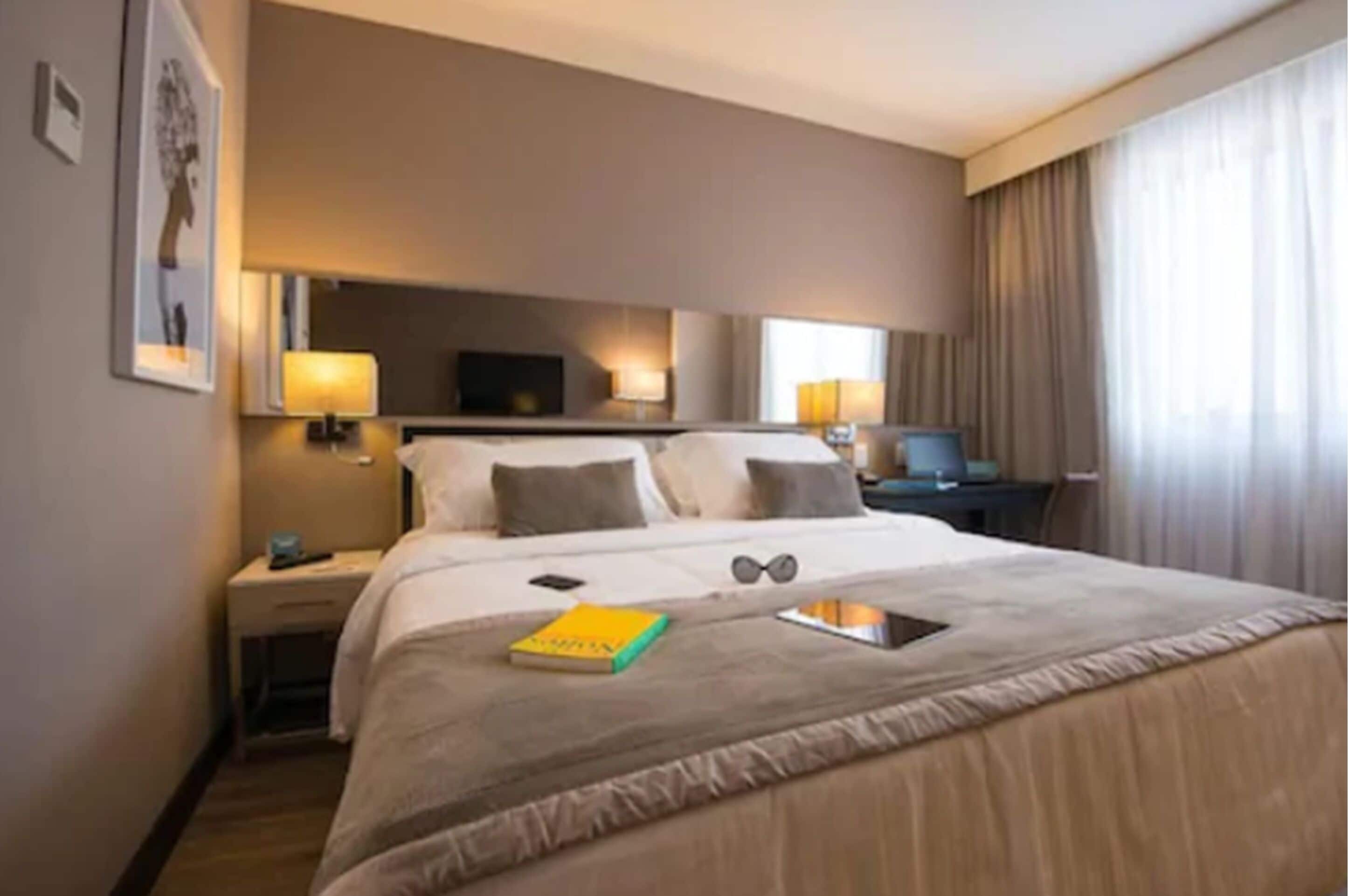 Une chambre d’hôtel avec un lit, des tables de chevet, un bureau et une grande fenêtre avec des rideaux.