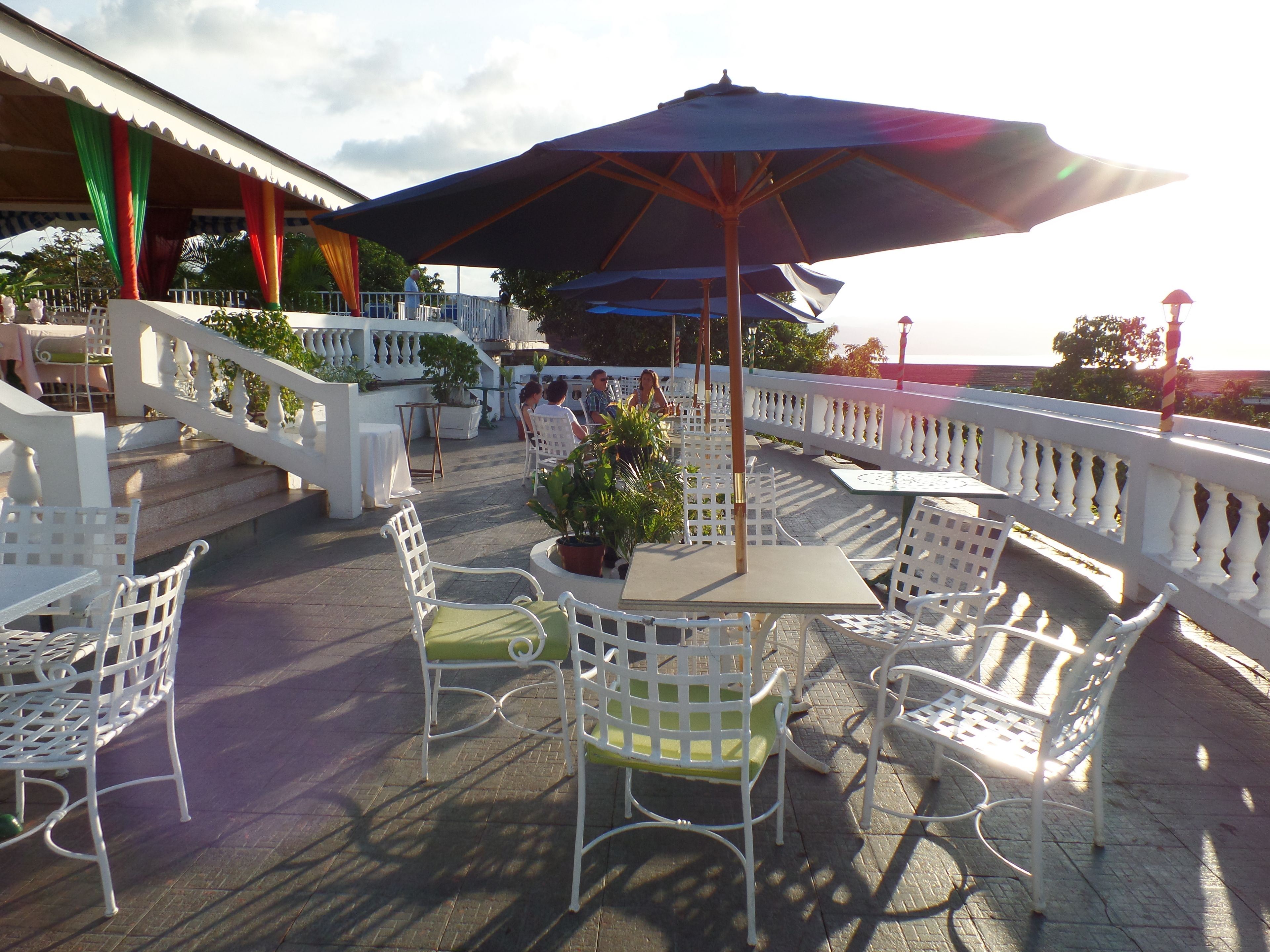 An outdoor seating area with white metal chairs and tables, a large umbrella, and a white railing.