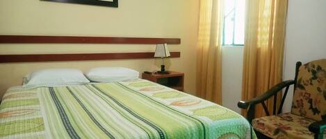 Habitación de hotel con cama, una silla, una mesita de noche, una lámpara y un cuadro en la pared.