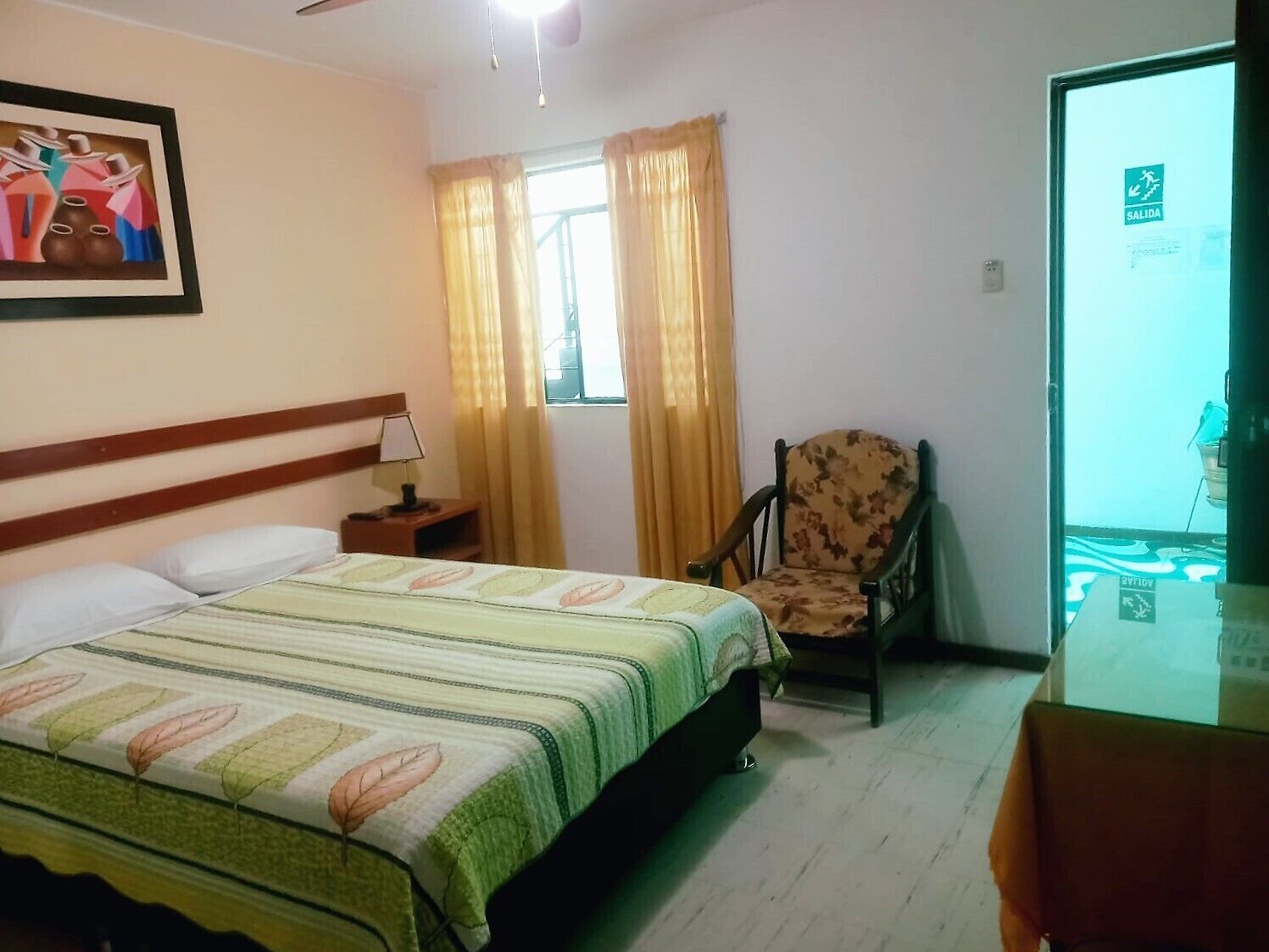 Una habitación de hotel con cama, una silla, una mesita de noche, un cuadro y una ventana con cortinas.