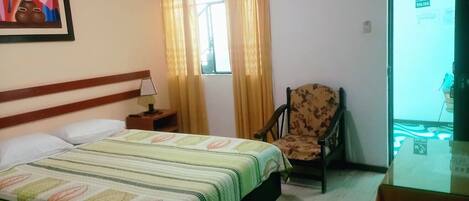 Una habitación de hotel con cama, una silla, una mesita de noche, un cuadro y una ventana con cortinas.