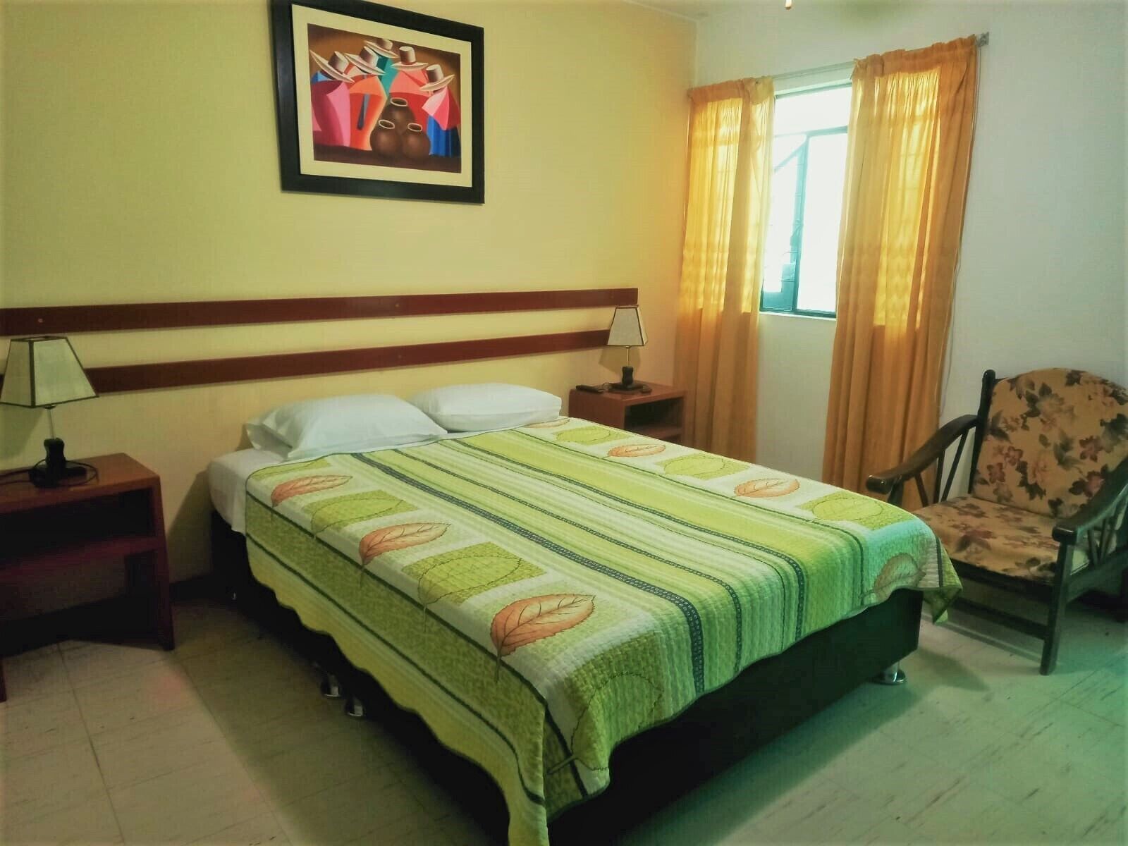 Habitación de hotel con cama, dos mesitas de noche, una silla, ventana con cortinas y un cuadro en la pared.