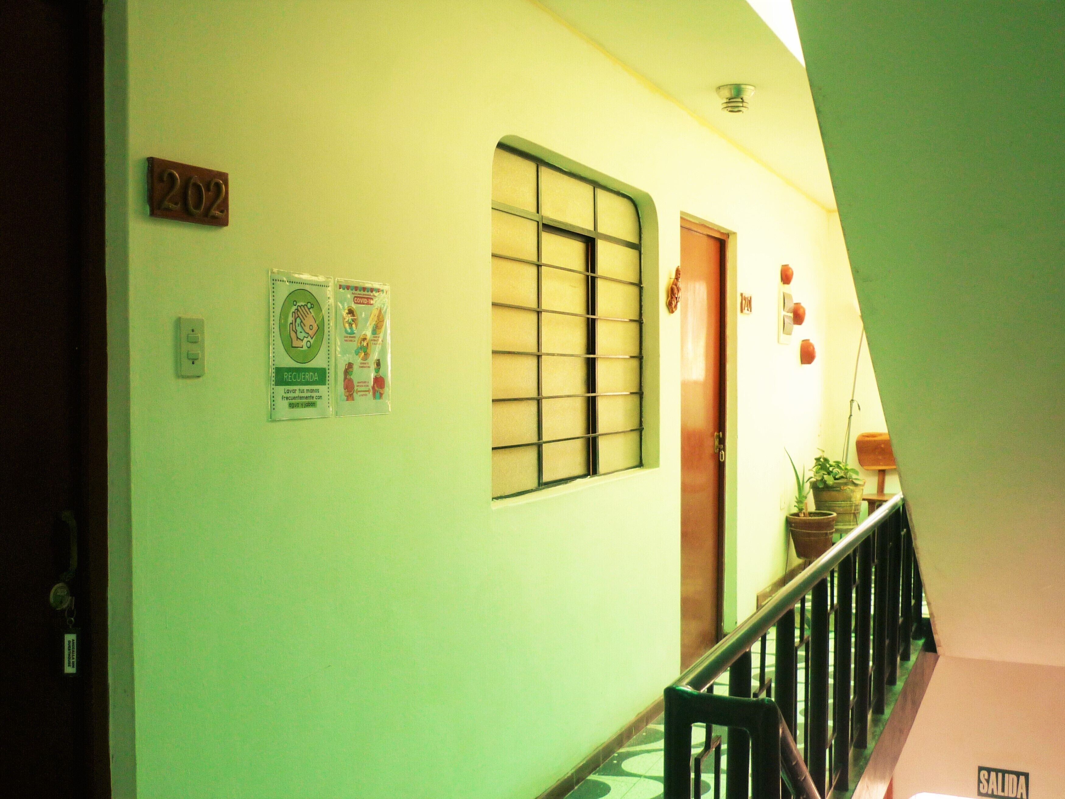 A hallway with a green wall, a door labeled '202', a window with bars, and a sign with health guidelines.