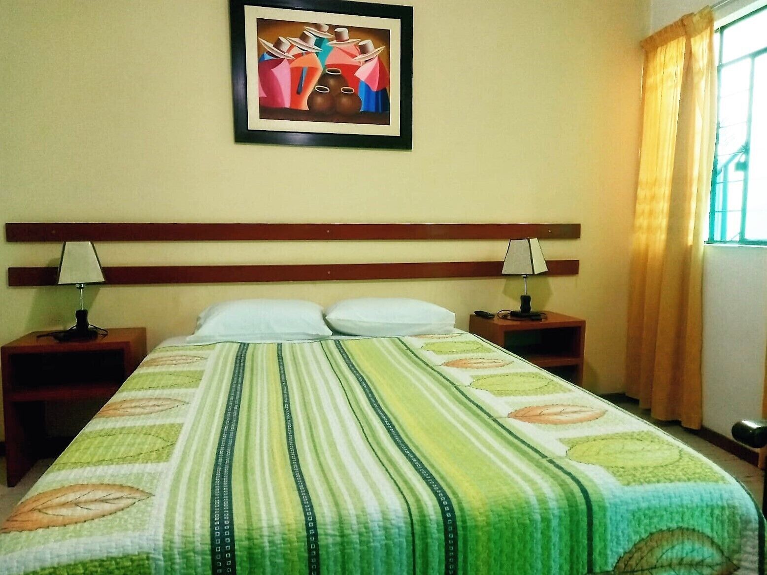 Habitación de hotel con cama, dos mesitas de noche con lámparas, un cuadro enmarcado en la pared y una ventana con cortinas.