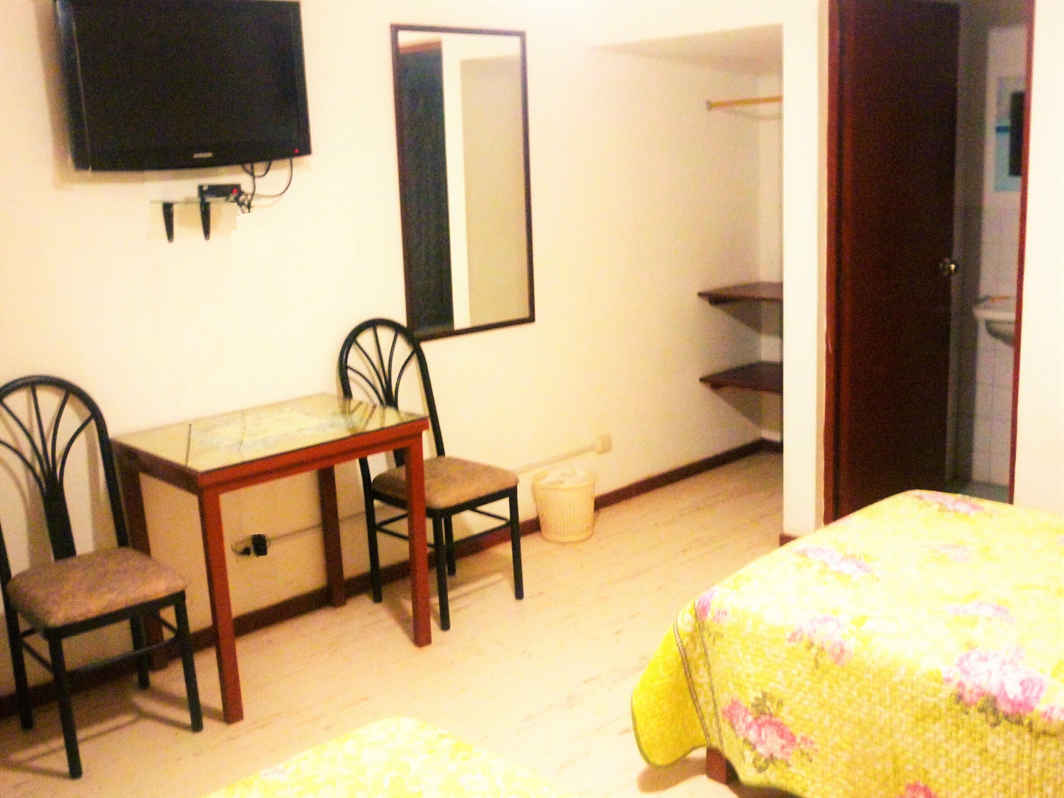 A small room with a wooden table, two chairs, a television mounted on the wall, and a bed with a floral bedspread.