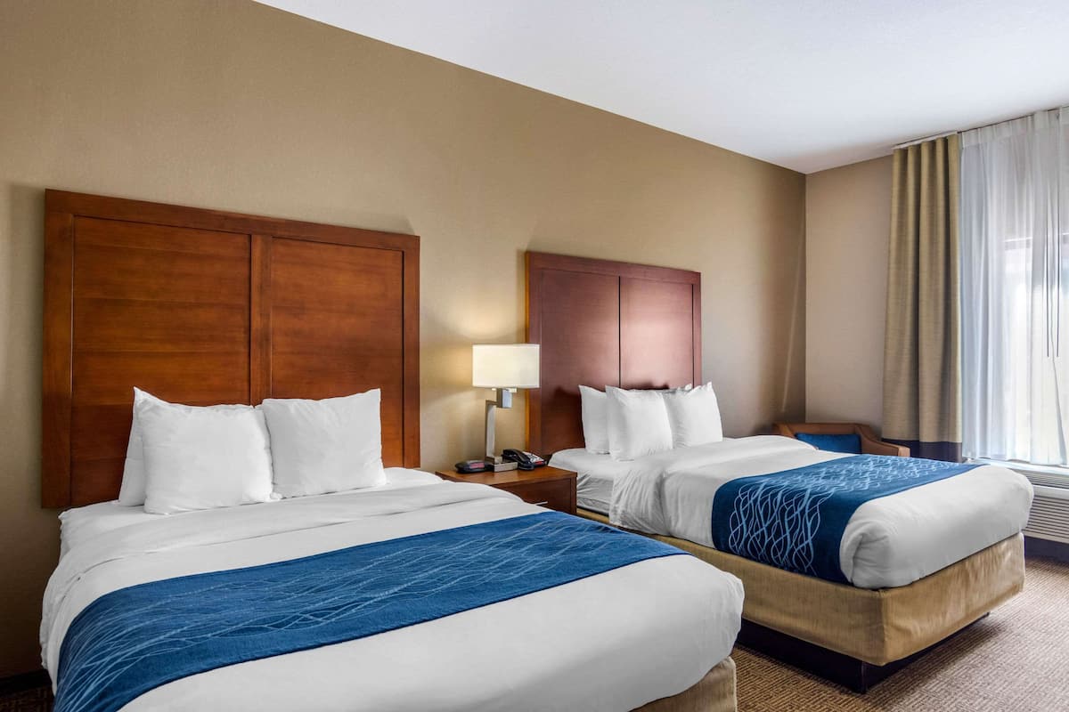 A hotel room with two beds, a wooden headboard, a bedside table with a lamp, and a window with curtains.