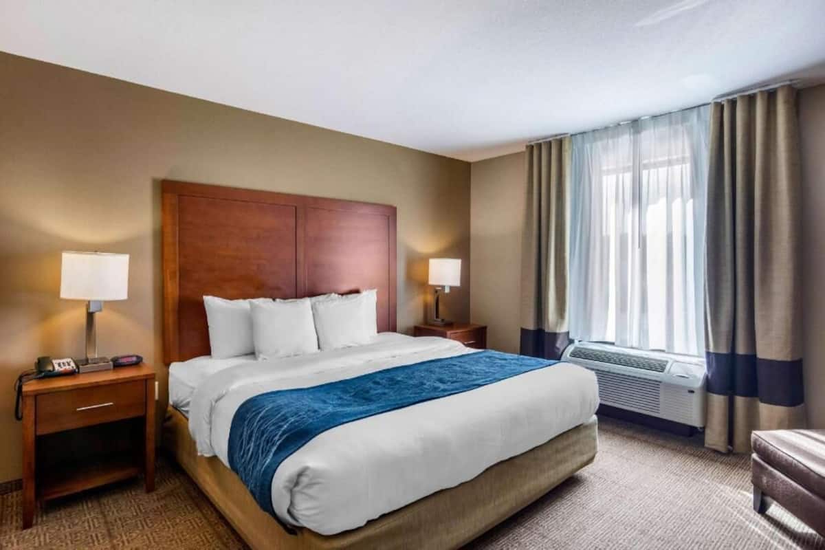 A hotel room with a bed, two bedside tables, a nightstand, a window with curtains, and an air conditioning unit.