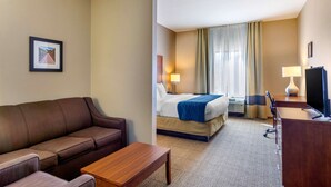 A hotel room with a brown leather sofa, a wooden coffee table, a bed with a blue blanket, a desk with a lamp, and a television. - Comfort Inn South Chesterfield - Colonial Heights (Colonial Heights)