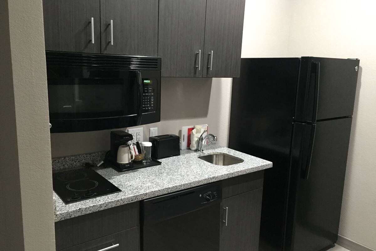 Una cocina compacta con gabinetes oscuros, un microondas negro, un refrigerador negro, una cafetera y un lavamanos.