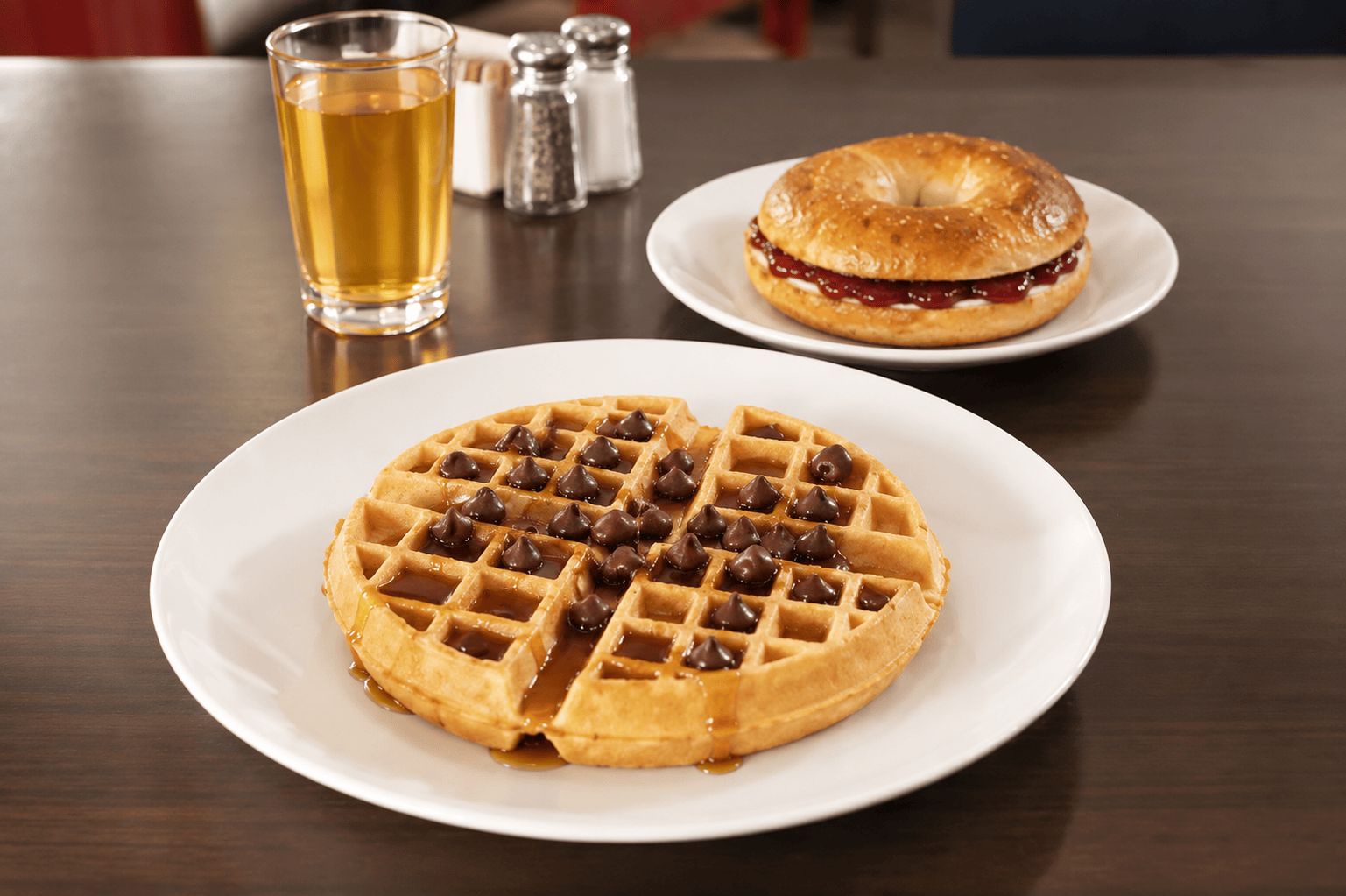 Una cialda con gocce di cioccolato e sciroppo su un piatto, un bicchiere di birra e un bagel con marmellata su un altro piatto.