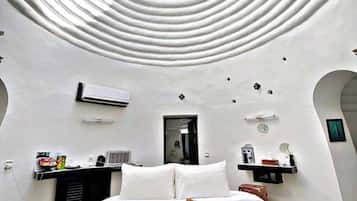 A modern bedroom with a circular skylight, a bed with white linens, a sun-shaped pillow, and a small bedside table with a mirror and a clock.