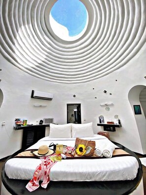 Un dormitorio moderno con un tragaluz circular, una cama con ropa de cama blanca, un almohadón con forma de sol y una mesita de noche con un espejo y un reloj.