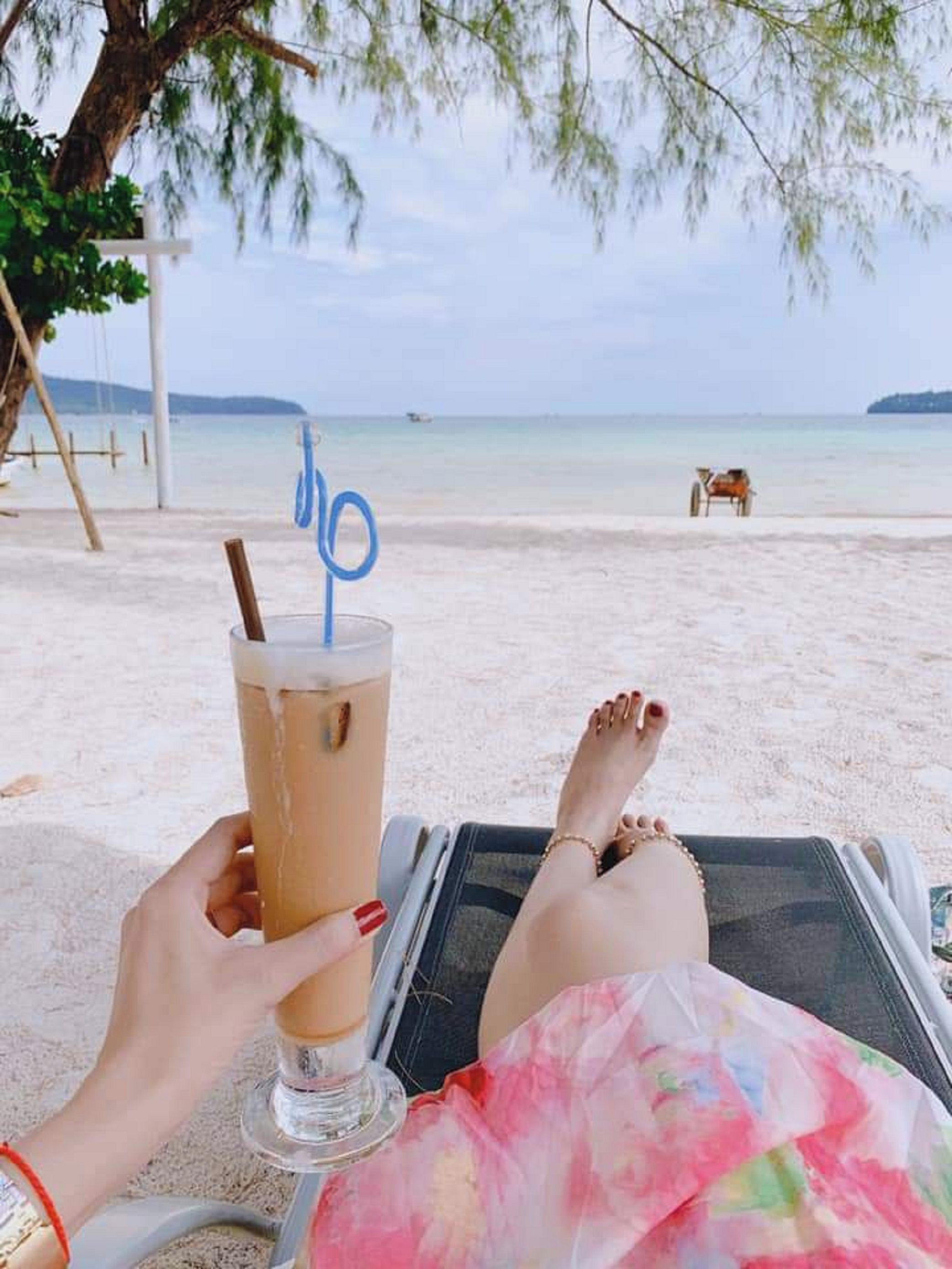 A person relaxing on a beach chair with a drink, a beach in the background, and a cart.