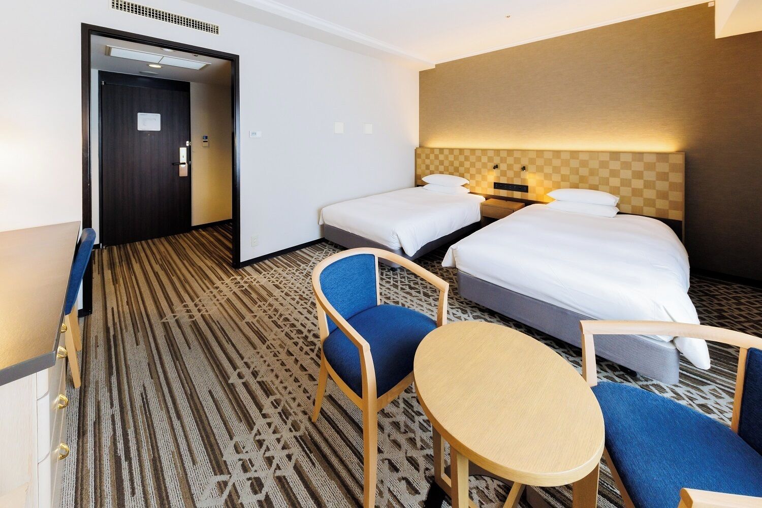 Une chambre d’hôtel avec deux lits, une petite table, deux chaises et une moquette à motifs.