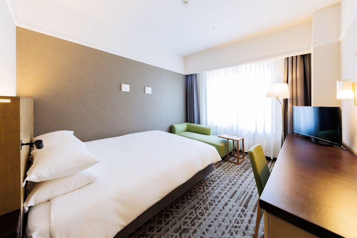 Una habitación de hotel con cama, escritorio, televisor y ventana con cortinas.