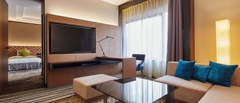 A modern hotel room with a sofa, a glass coffee table, a flat-screen TV, a desk, and a bed.