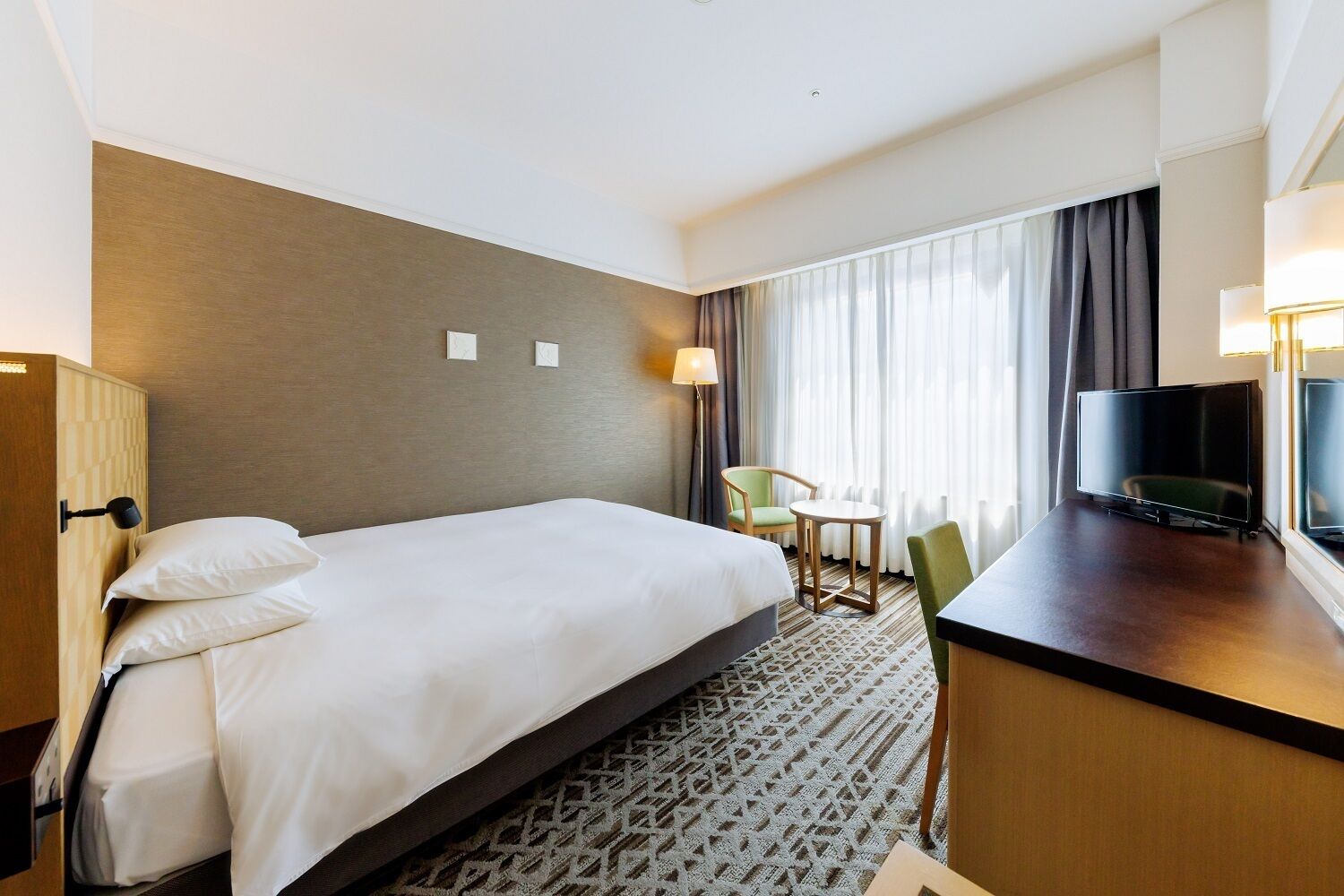Ein Hotelzimmer mit einem großen Bett, einem Schreibtisch mit Fernseher, einem Sessel und einem Fenster mit Vorhängen.