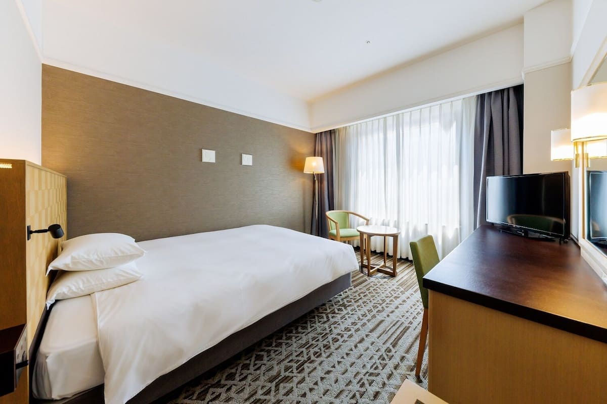 Habitación de hotel con una cama grande, un escritorio con televisor, una silla y una ventana con cortinas.
