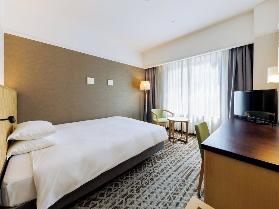 A hotel room with a large bed, a desk with a flat-screen TV, a chair, and a small table.